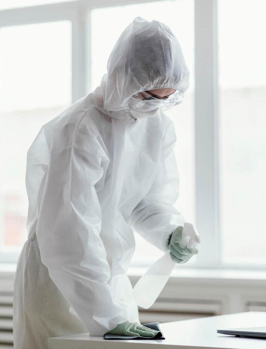 Kobieta ubrana w specjalną odzież ochronną, okulary, maseczkę i rękawiczki czyści biurko za pomocą środka chemicznego.